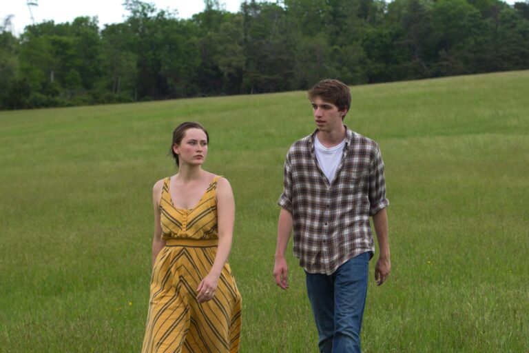Couple walking in a grassy field; woman in yellow dress, man in plaid shirt.