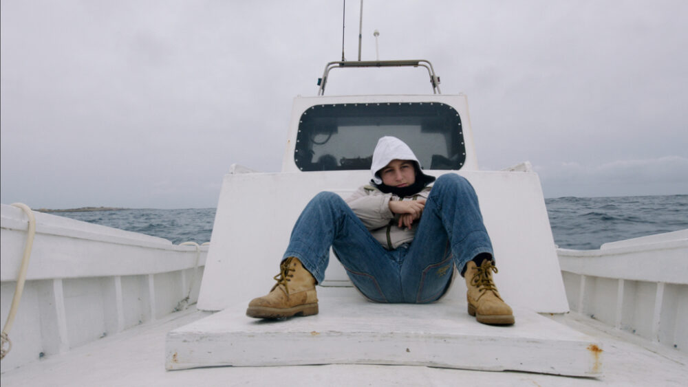 Boy on a boat at sea, wearing a hooded jacket and boots.