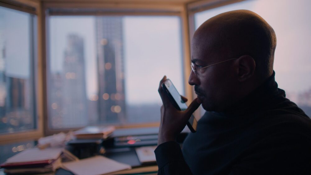 Man in turtleneck contemplates phone, city skyline backdrop.