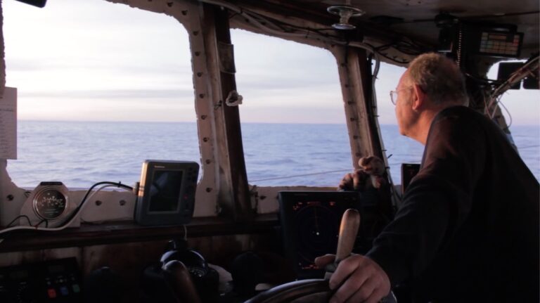 Fisherman at the helm of a boat, looking out at the ocean.