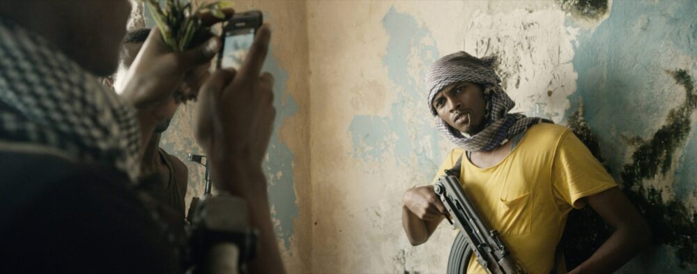Man with rifle in "Fishing Without Nets" film being photographed by a person with a phone.