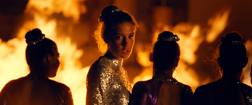 Woman in sequined dress, possibly from "The Five Devils" film, surrounded by dancers against a fiery backdrop.