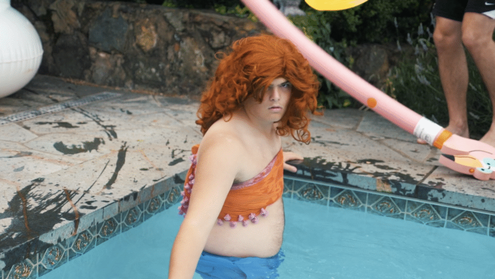 Person in pool wearing orange top and red wig.
