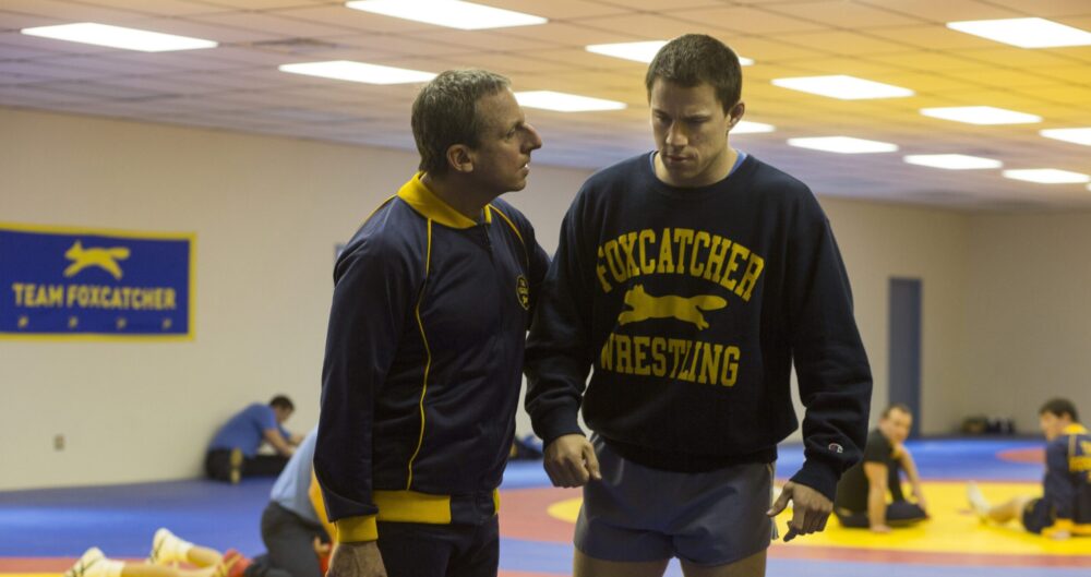 Channing Tatum in Foxcatcher with coach, wrestling practice in background.