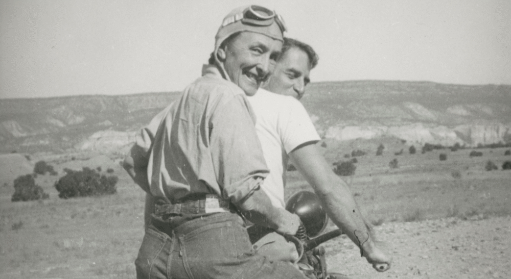 Georgia O'Keeffe on a motorcycle, wearing goggles, with a man in a desert landscape