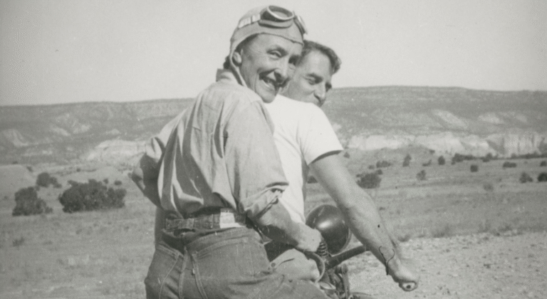 Georgia O'Keeffe on a motorcycle, wearing goggles, with a man in a desert landscape