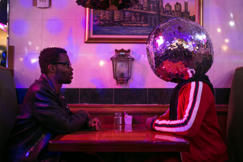 Man in leather jacket facing person with disco ball head in unique restaurant setting.