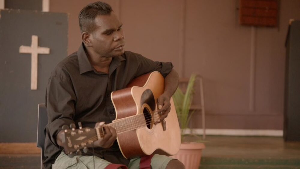 Gurrumul, an Aboriginal musician, playing guitar in front of a cross.