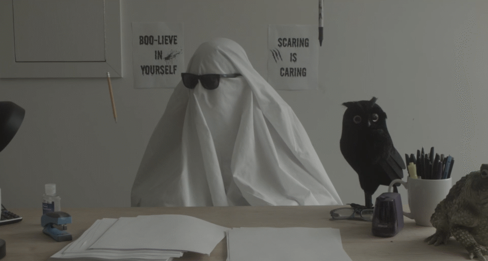 Ghost at desk with "Boo-lieve in Yourself" and "Scaring is Caring" signs.