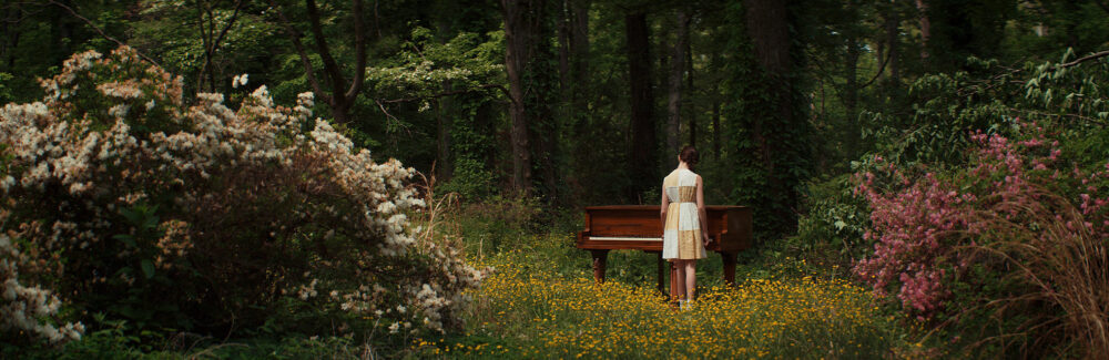 Woman stands at a piano in a forest clearing with flowers.