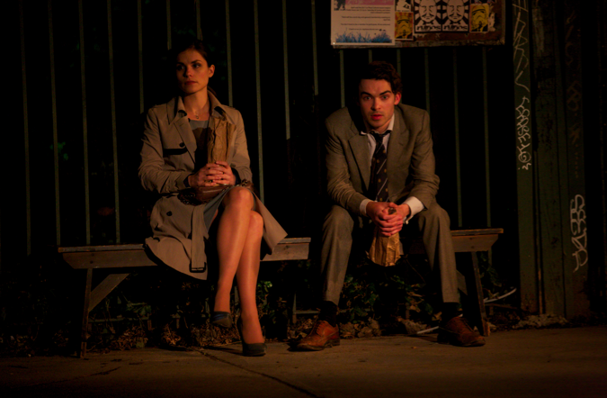 Couple sitting on park bench at night, each holding a brown paper bag.