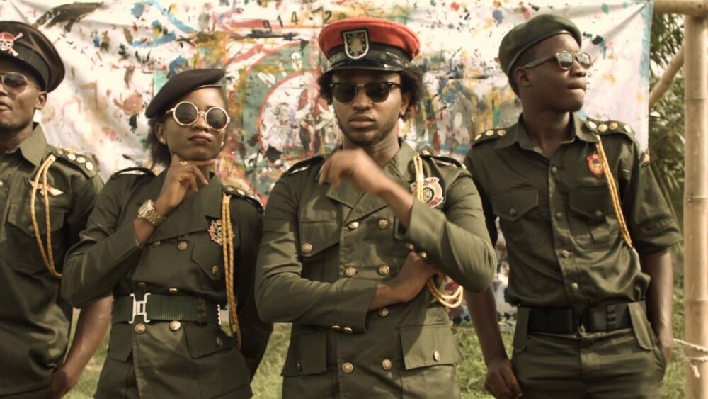 Four people in green military-style uniforms and sunglasses pose in front of a colorful backdrop.