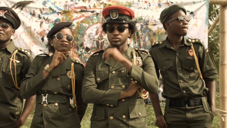 Four people in green military-style uniforms and sunglasses pose in front of a colorful backdrop.