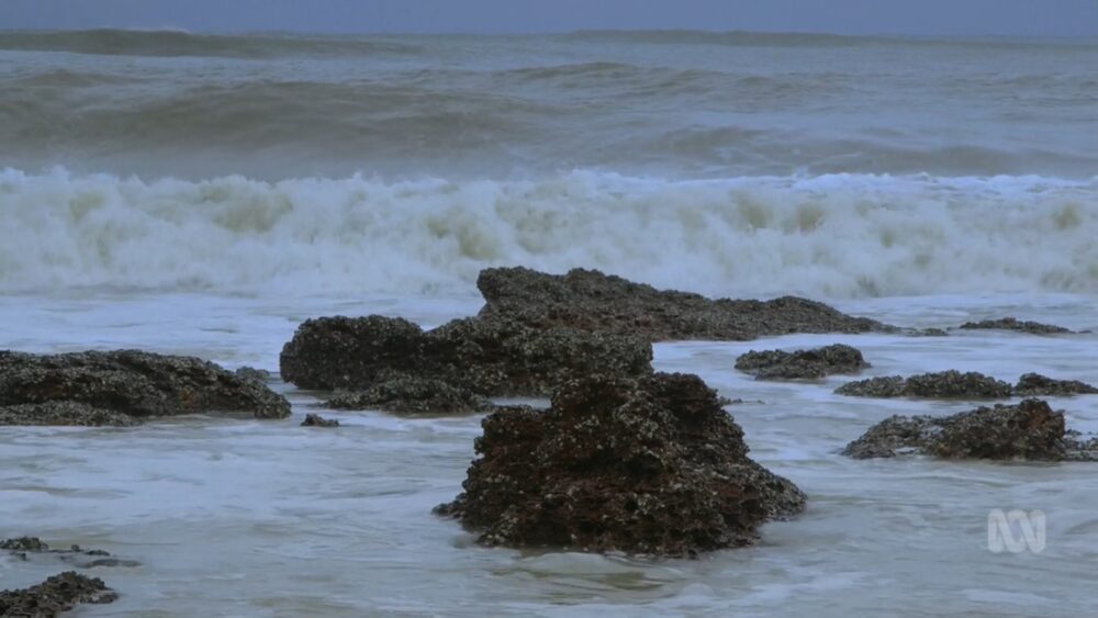 Rough ocean waves crash against dark, rocky outcrops along the shore under a cloudy sky.