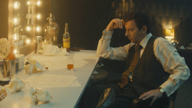 Pensive man in vest sits at dressing table with script pages and a drink.