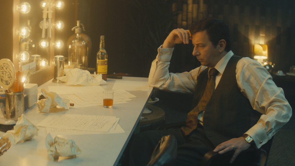 Pensive man in vest sits at dressing table with script pages and a drink.