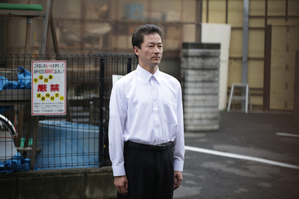 Japanese man in a white shirt and black pants standing outdoors, near a sign with Japanese text.