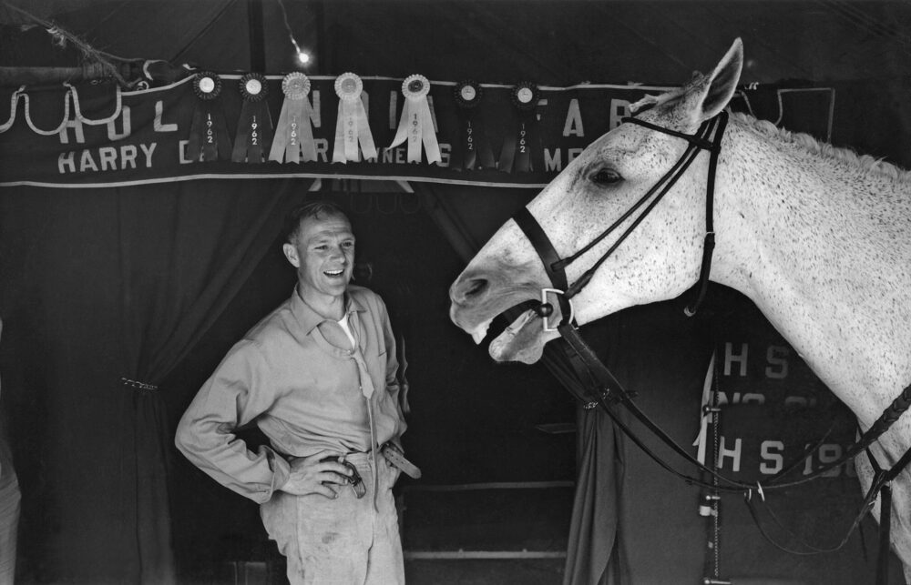 Harry de Leyer with Snowman, a gray horse, at a show. Ribbons with "1962" adorn the tent.