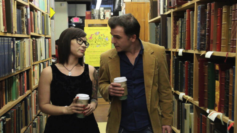 Couple with coffee walks through a bookstore, browsing shelves of vintage books.