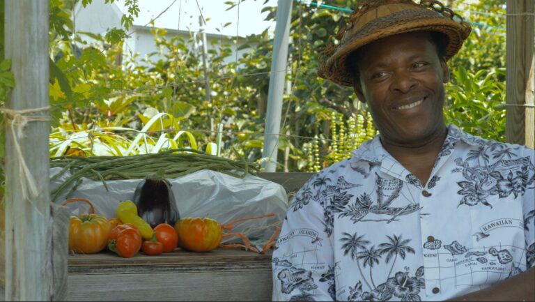 Smiling Black farmer with fresh produce: tomatoes, eggplant, and long beans, wearing a Hawaiian shirt and straw hat.