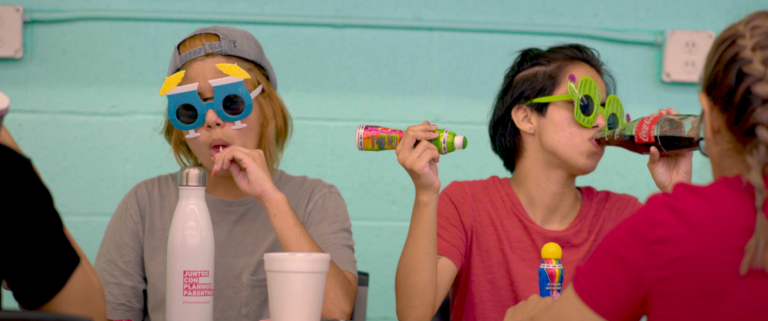 Friends with novelty glasses drinking soda at a table. "Juntos Crecemos Planned Parenthood" on water bottle.
