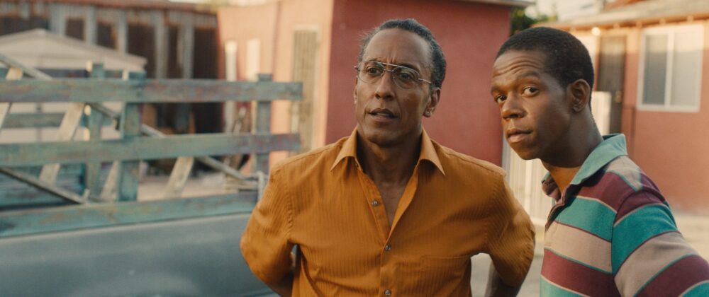 Two men, one in an orange shirt and glasses, stand outside in Hunter Gatherer.