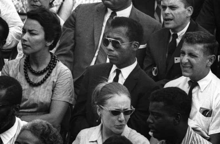 James Baldwin in sunglasses at a public event, thoughtful expression.