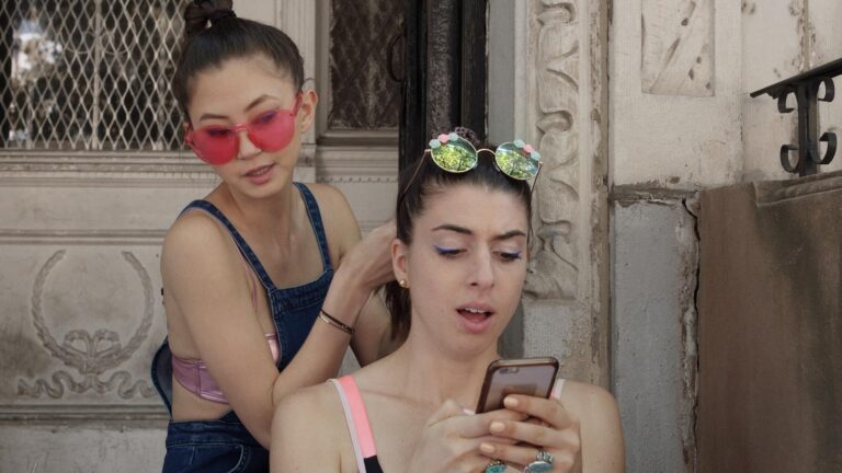 Two young women, one fixing the other's hair while she looks at her phone outdoors.