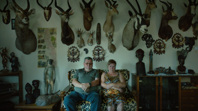 Couple seated amidst taxidermied animal heads in "In the Basement".