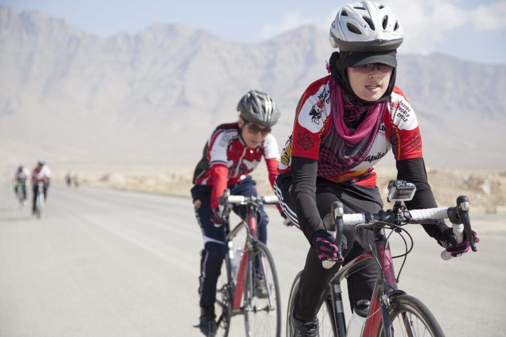 Women cycling in "Afghan Cycles" race, wearing helmets and jerseys, with mountains in the background.