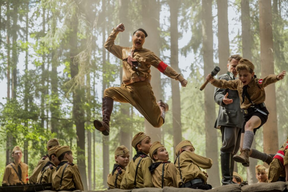 Jojo Rabbit image: Taika Waititi as Hitler jumping with Hitler Youth members in the forest.