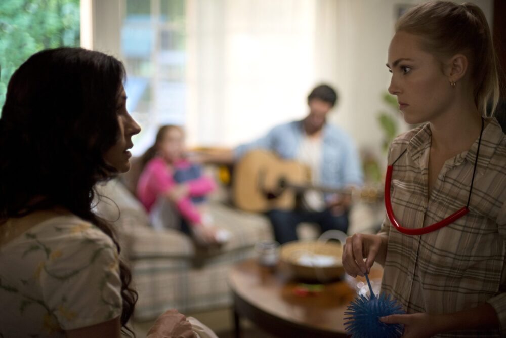Two women talking indoors, man with guitar in background. Jack of the Red Hearts scene.