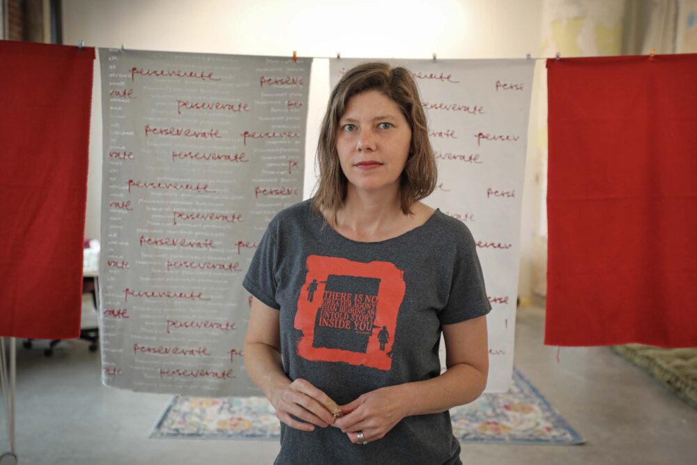 Artist Joann Self Selvidge stands before her art, wearing a shirt that reads: "There is no greater agony than bearing an untold story inside you.