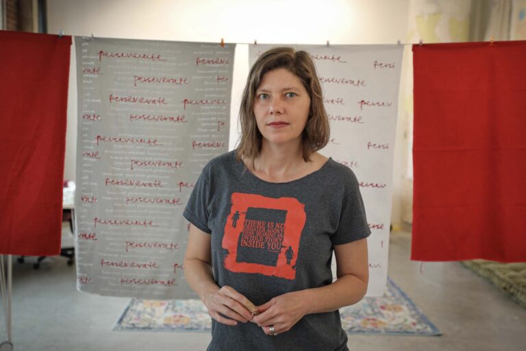 Artist Joann Self Selvidge stands before her art, wearing a shirt that reads: "There is no greater agony than bearing an untold story inside you.