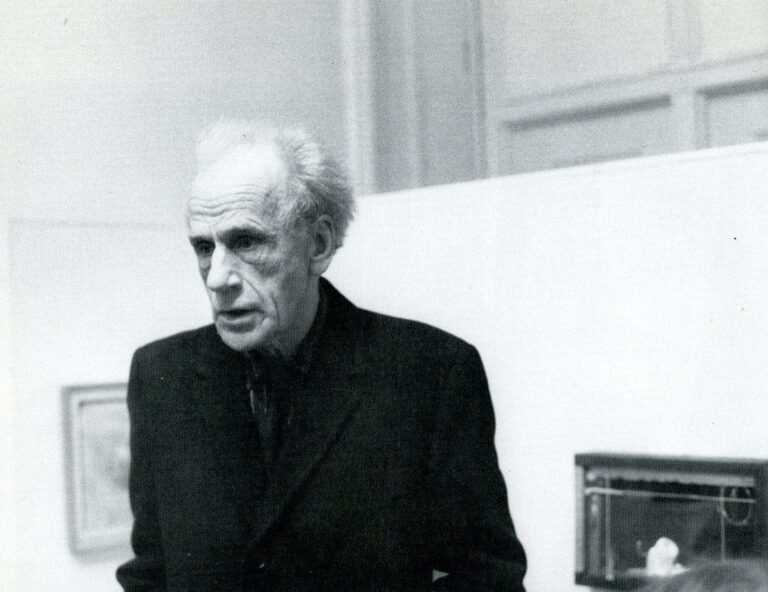 Joseph Cornell at gallery, wearing dark suit. Boxes and films in background.
