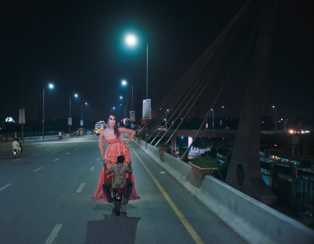 Joyland: Transgender woman in red dress stands behind man on motorcycle at night on bridge.