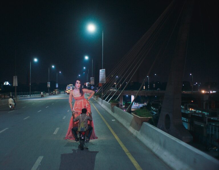 Joyland: Transgender woman in red dress stands behind man on motorcycle at night on bridge.