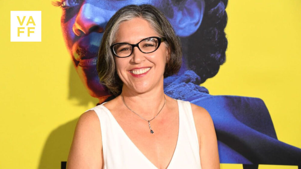 Nicole Kassell smiles wearing glasses and a white dress in front of a colorful mural.