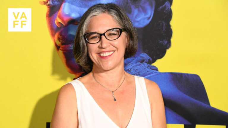 Nicole Kassell smiles wearing glasses and a white dress in front of a colorful mural.