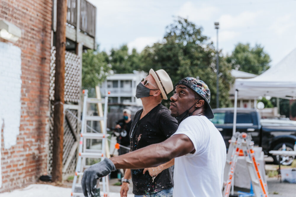 Two men mending walls, looking up at a mural project. Ladders and painting equipment are visible.
