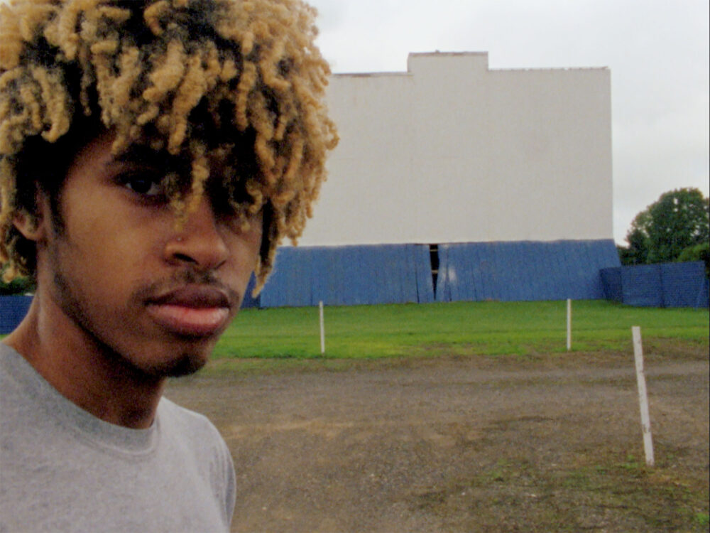 Portrait of a man with blonde dreadlocks in front of a drive-in movie screen.