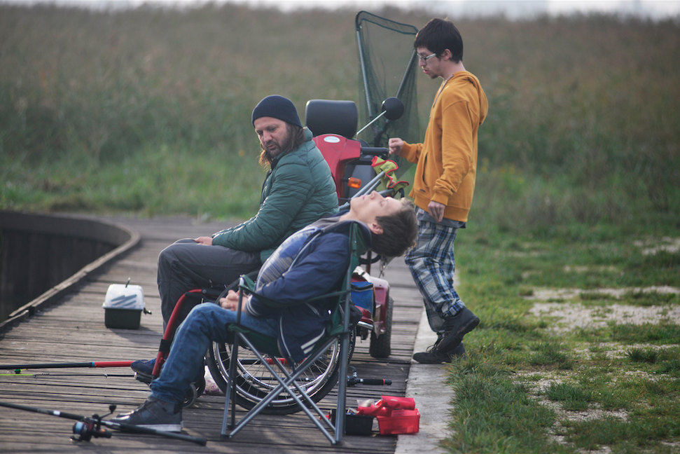 Three people fishing by a dock, one resting in a wheelchair near a small red vehicle.