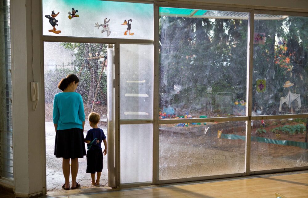 A kindergarten teacher and child look out at a rainy playground through a glass door.