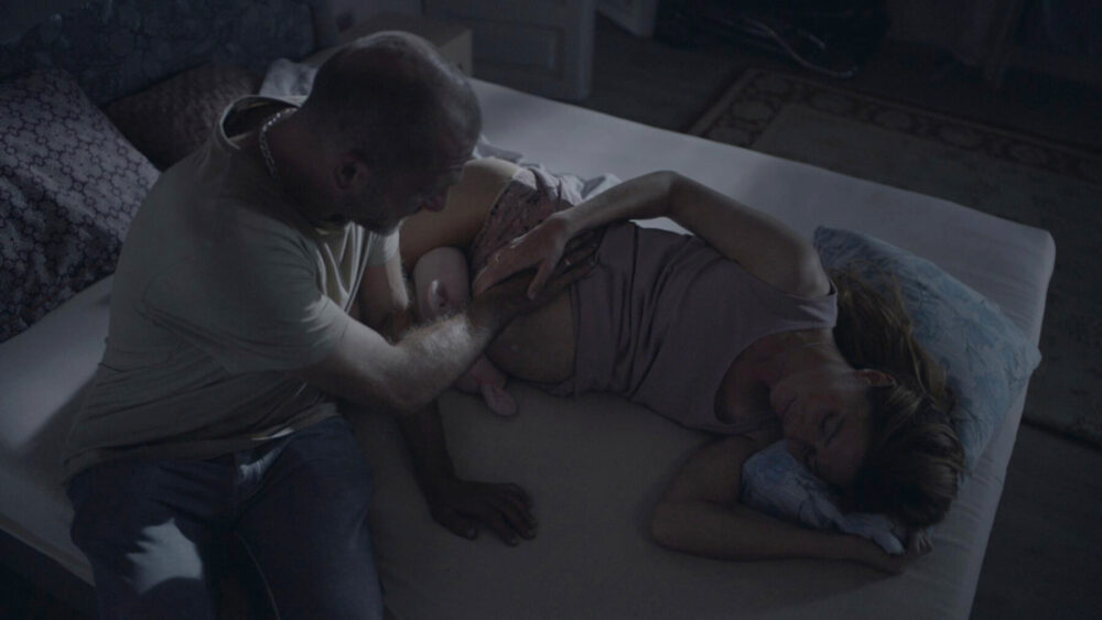 Man touching a sleeping woman on a bed. The scene has a somber mood.