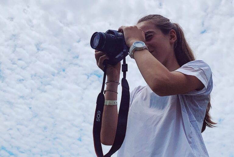 Photographer with Canon EOS camera against a cloudy sky, wearing a white t-shirt.