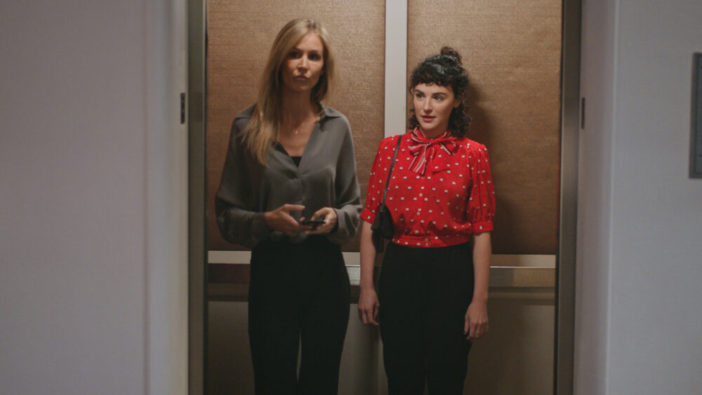 Two women in office attire stand in an elevator, one blonde and one with dark curly hair.