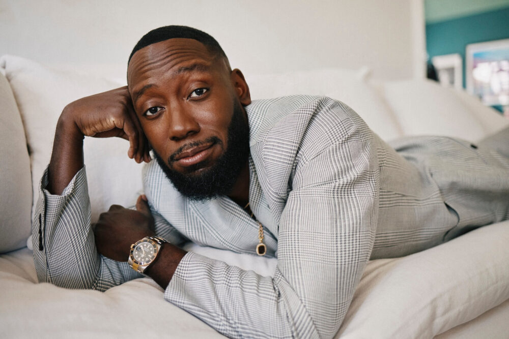 Lamorne Morris lying on a white couch, wearing a gray plaid suit and gold watch.