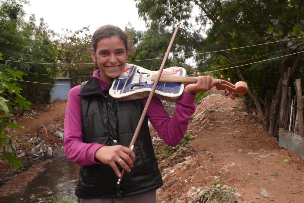 Young woman smiles, playing a violin made from recycled materials near a landfill. Landfill Harmonic project.