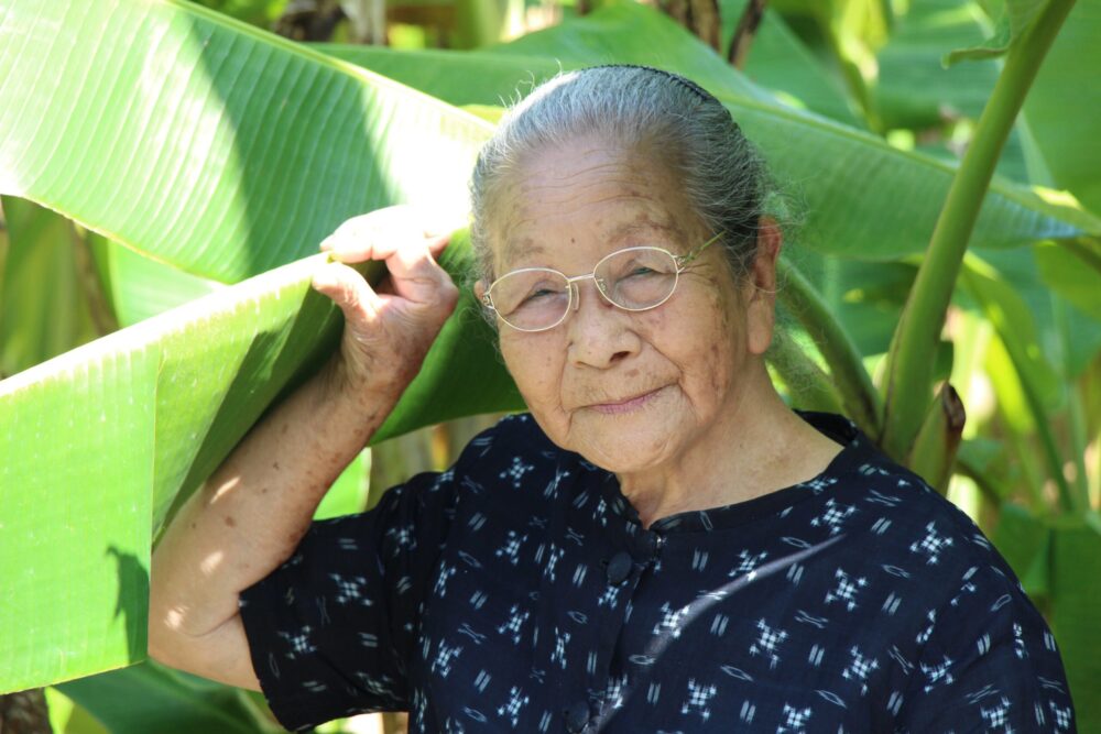 Elderly Asian woman with glasses stands by tropical banana leaves, part of Landscapes of Longevity.