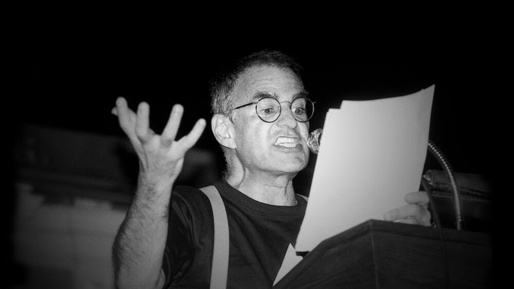 A black and white photo of Larry Kramer passionately speaking at a podium, holding a paper.